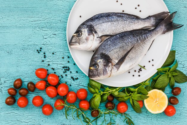 Dwie świeże ryby dorado na białym talerzu i warzywa na niebieskim stole. Widok z góry, miejsce na kopię.