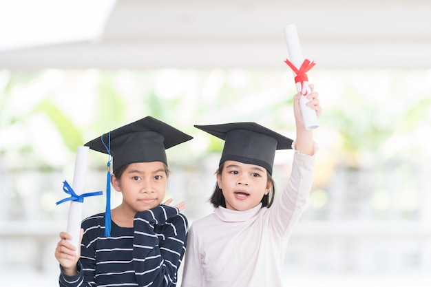 Dwie szczęśliwe azjatyckie absolwentki szkoły żeńskiej z czapką dyplomową posiadają zwinięty certyfikat. Graduation Celebration Concept Stock Photo