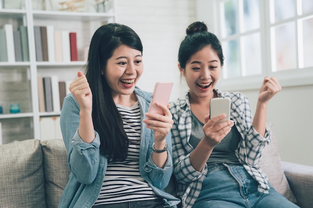 dwie szczęśliwe azjatyckie koleżanki siedząc na kanapie w domu w salonie, uśmiechając się, śmiejąc się z wesołą twarzą, wygrywając gest rąk patrząc na smartfona. młode kobiety za pomocą telefonu komórkowego w pomieszczeniu radosne.