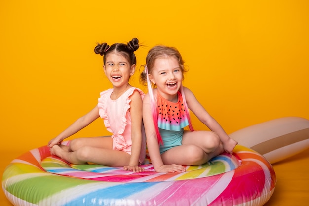 Dwie szczęśliwe dziewczynki w strojach kąpielowych siedzi na kolorowym nadmuchiwanym materacu lizak na żółtej powierzchni.