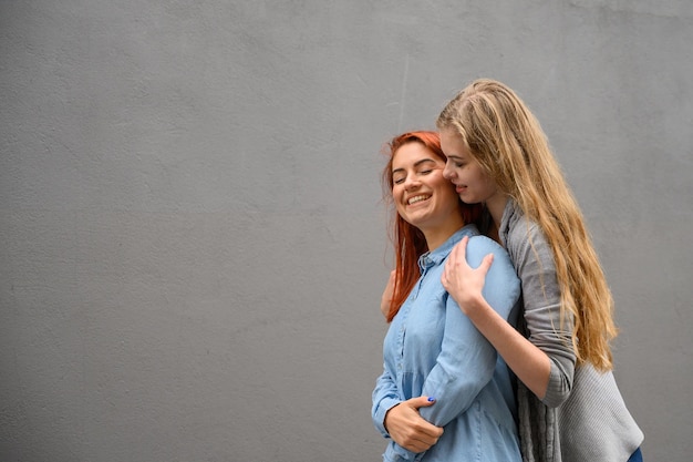 Dwie szczęśliwe dziewczyny przytulają się na tle szarej ściany Delikatne uściski żeńskiej pary lesbijek LGBT Małżeństwo osób tej samej płci