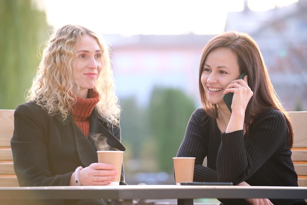 Dwie szczęśliwe koleżanki bawią się razem w kawiarni ulicznej, śmiejąc się i rozmawiając przez telefon komórkowy