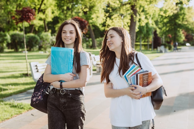Dwie szczęśliwe uśmiechnięte dziewczyny studentki chodzą i rozmawiają ze sobą na terenie kampusu w ciepły słoneczny dzień. Miękka selektywna ostrość.