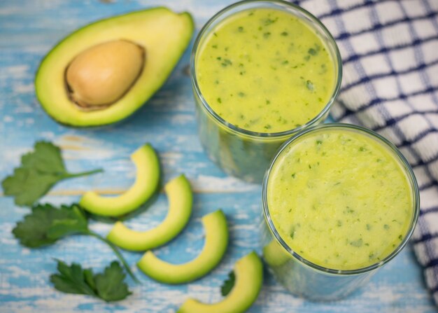 Zdjęcie dwie szklane filiżanki z koktajlem z awokado, banana, kiwi i ziół na niebieskim drewnianym stole. dieta wegetariańska.