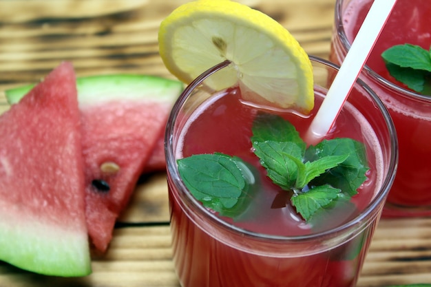 Dwie szklanki soku z arbuza ze słomką ozdobioną listkami mięty i cytryny stoją na drewnianym stole. Pyszny i zdrowy sok.