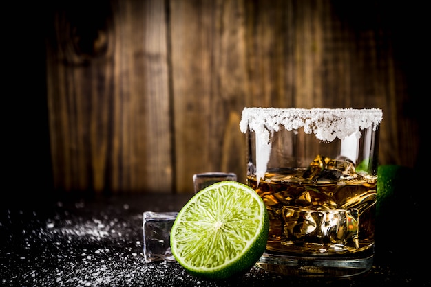 Dwie szklanki tequili na ciemnym stole, z kostkami lodu, solą i limonką