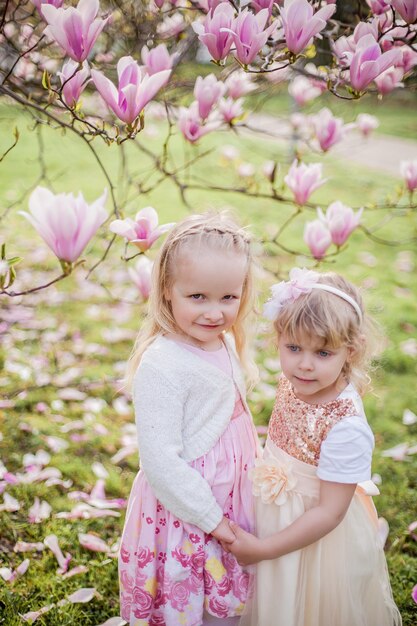 Dwie Urocze Blondynki W Wieku 3 Lat Bawią Się W Parku W Pobliżu Kwitnącej Magnolii. Picie Herbaty. Wielkanoc. Wiosna.