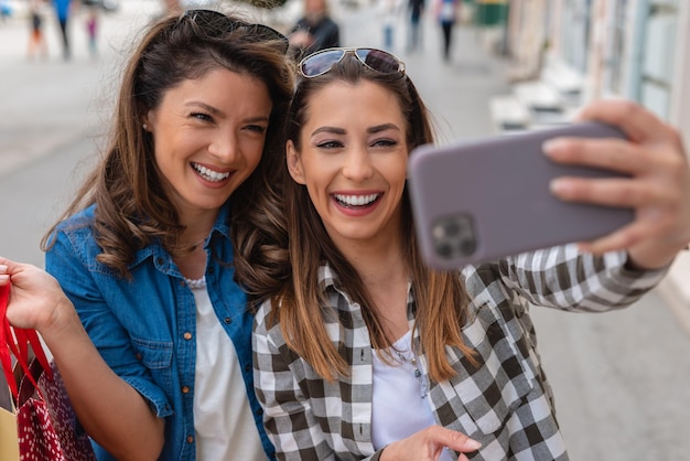 Dwie urocze uśmiechnięte kobiety robiące selfie na smartfonie trzymające torby na zakupy