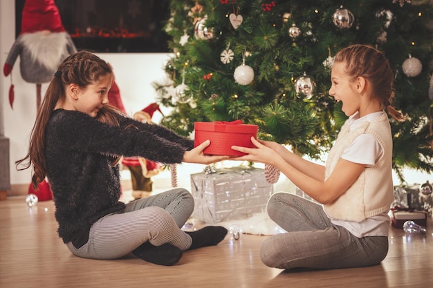 Dwie uśmiechnięte dziewczynki siedzą na podłodze przy choince. Jeden daje drugiemu prezent w czerwonym pudełku.
