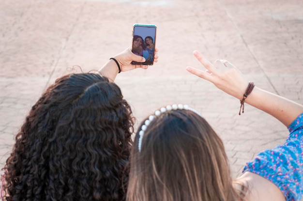 Dwie uśmiechnięte koleżanki nastolatka robiące selfie i wskazujące palce w górę Zbliżenie dwóch uśmiechniętych nastoletnich koleżanek siedzących przy selfie