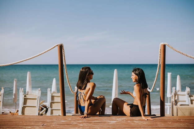 Dwie uśmiechnięte młode kobiety w bikini siedzące i cieszące się wakacjami na plaży