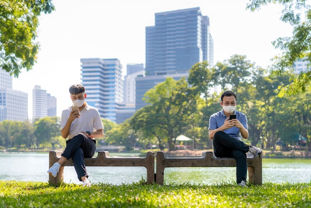 Dwóch azjatyckich młodych ludzi na czacie w smartfonie i noszących maskę w odległości 6 stóp chroni przed wirusami COVID-19 w celu zdystansowania się w społeczeństwie przed ryzykiem infekcji w parku