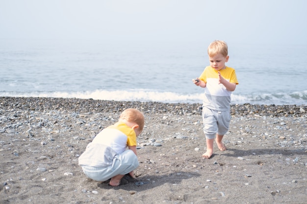 Dwóch braci bliźniaków młodszy chłopiec bawi się skałami i piaskiem na plaży w słoneczny dzień.