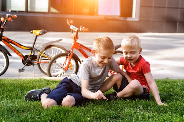 Dwóch chłopców gra siedząc na trawie. Odpoczynek po jeździe na rowerze, w tle Rowery