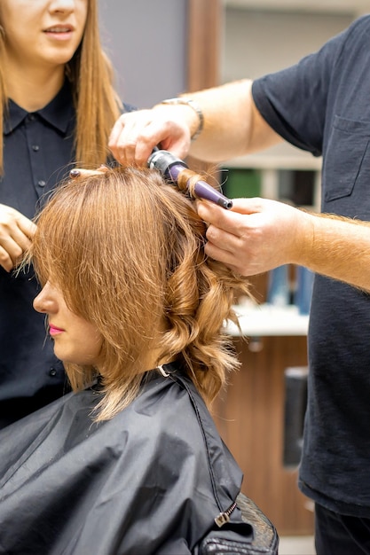 Dwóch fryzjerów robi loki fryzurę długich brązowych włosów za pomocą lokówki w salonie fryzjerskim