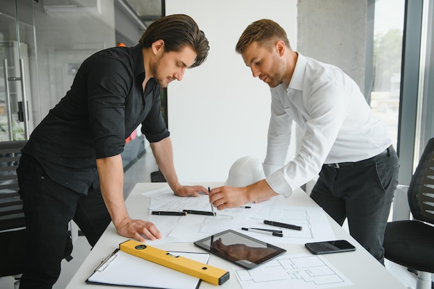 Dwóch inżynierów pracujących razem i korzystających z cyfrowego tabletu wyglądającego na plan i analizę z planem architektonicznym na biurku