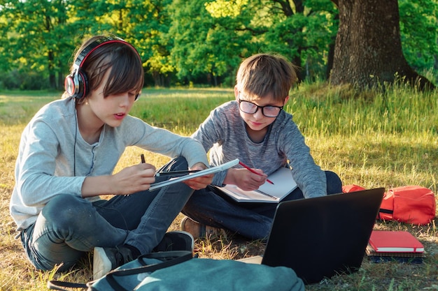 Dwóch kolegów z klasy studiujących siedzących na ziemi w parku przed laptopem i odrabiających lekcje w zeszytach ćwiczeń Chłopiec w okularach uważnie obserwujących ucznia w słuchawkach, który czyta zadanie z notatnika