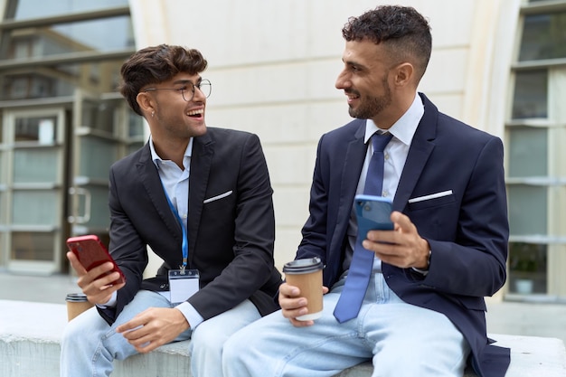 Dwóch latynoskich pracowników biznesowych używających smartfonów do picia kawy na ulicy