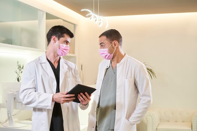 Dwóch lekarzy z maską komentujących wyniki z tabletką w dłoni. Poradnia ginekologiczna, stomatologiczna lub estetyczna. Pojęcie medyczne.