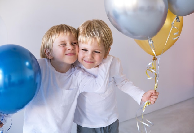 Dwóch małych blondynów uśmiechających się chłopców bliźniaków obejmujących się, trzymających balony, świętujących urodziny.