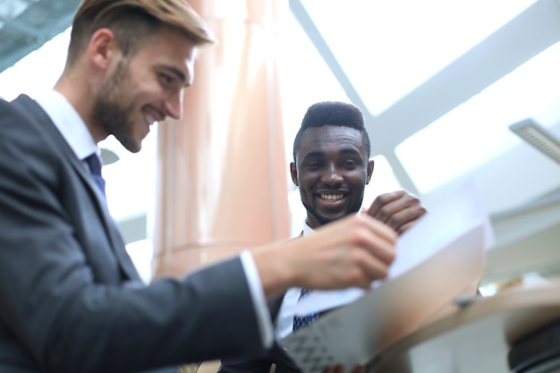 Zdjęcie dwóch międzynarodowych młodych biznesmenów w garniturze, omawiając coś i wskazując papier.