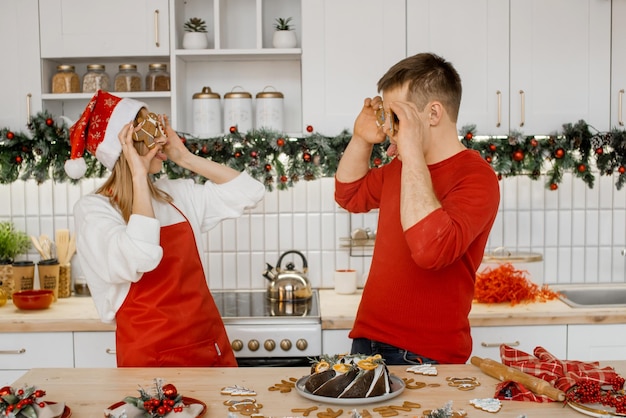 Dwóch miłych dziecinnych, wesołych, pozytywnych małżonków, mąż, żona, bawi się w kuchni pokazując sobie języki i zakrywając oczy piernikowymi ciasteczkami