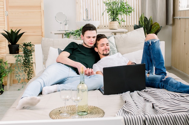 Dwóch młodych gejów leżących na łóżku i korzystających z laptopa. Przystojni homoseksualiści spędzają czas razem. Para homoseksualna, geje. Obsługuje streamować film na laptopie w łóżku