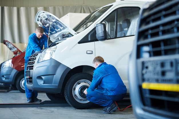 Dwóch młodych mechaników w mundurach robi przegląd techniczny samochodu lub naprawia jego silnik i koła w serwisie