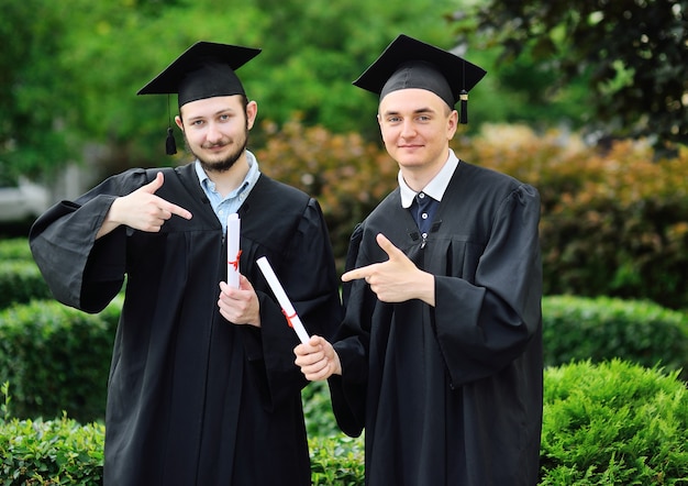 Dwóch młodych mężczyzn-absolwentów uczelni w togach i kwadratowych kapeluszach z radością odbiera dyplom.