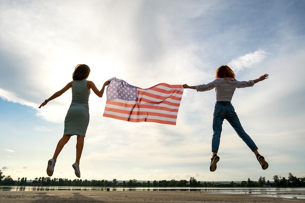 Dwóch młodych przyjaciół kobiet z flagą narodową USA, skoki razem na zewnątrz na brzegu jeziora.