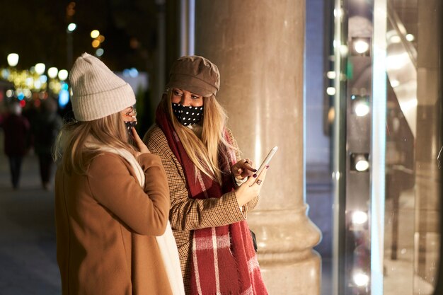 Dwóch Młodych Przyjaciół Patrząc W Wystawę Sklepową W Mieście Nocą. Jedna Z Nich Pokazuje Przyjaciółce Coś Na Jej Smartfonie.