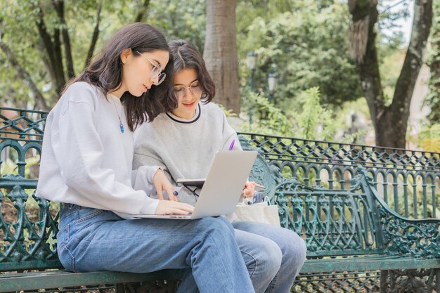 Dwóch młodych uczniów w szkole siedzi na ławce i wykonuje pracę szkolną z laptopem i notatnikiem.