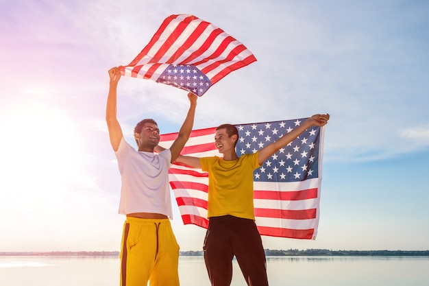 Dwóch młodych, wesołych i roześmianych przyjaciół Afroamerykanów i Kaukazu, dumnie trzymających amerykańską flagę na tle błękitnego nieba o zachodzie słońca w lecie