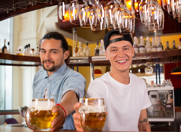 Dwóch młodych wesołych szczęśliwych barmanów patrząc na kamery i oferując piwo klientom. Radosni barmani są szczęśliwi, że są w pracy, mają miły dzień, cieszą się, że mogą służyć ludziom koncepcji.