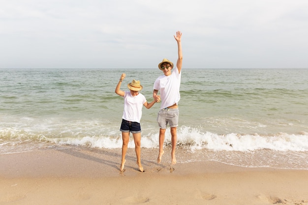 Dwóch nastolatków ubranych w białe T-shirty i szorty podskakuje trzymając się za ręce na morskiej plaży