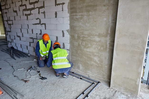 Dwóch pracowników lub budowniczych w odzieży ochronnej i pomarańczowych kaskach montujących plastikowe rury przy użyciu nowoczesnych narzędzi w mieszkaniu budowanego budynku