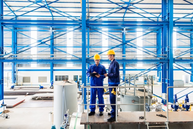 Dwóch pracowników w dużym warsztacie metalowym lub fabryce sprawdzających pracę stojącą na dużej maszynie