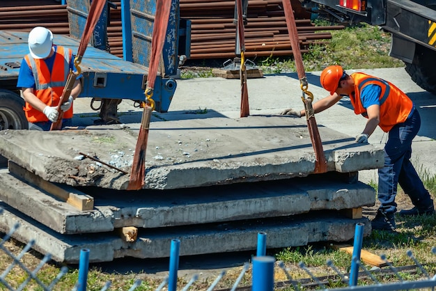 Dwóch procarzy rozładowuje betonowe płyty na ulicy w letni dzień Pracownicy w hełmach i kamizelkach budowlanych wyrzucają ładunek