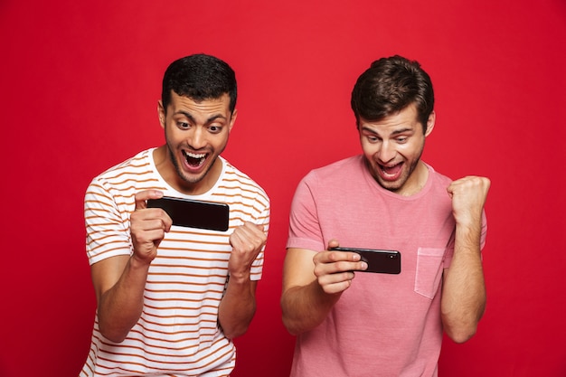 Dwóch przyjaciół wesołych mężczyzn stojących na białym tle nad czerwoną ścianą, grając w gry na telefonach komórkowych, świętując