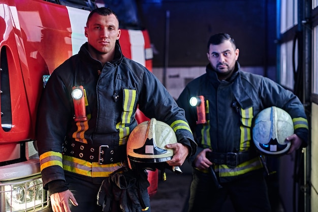 Dwóch strażaków w mundurach ochronnych stojących obok wozu strażackiego w garażu straży pożarnej.