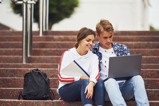 Dwóch studentów siedzących na schodach na zewnątrz z laptopem w ciągu dnia.