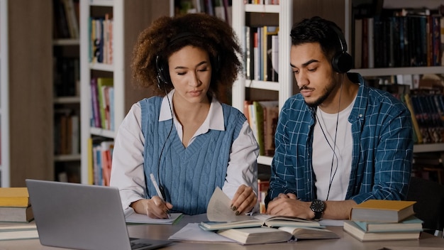 Dwóch studentów siedzi przy biurku w bibliotece uniwersyteckiej, słucha nauczyciela w słuchawkach na laptopie, uczy się pisać.