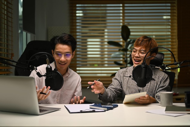 Dwóch szczęśliwych mężczyzn za pomocą laptopa i mikrofonu przesyłających strumieniowo podcast audio w domowym studiu Podcasty rozrywkowe i koncepcja technologii