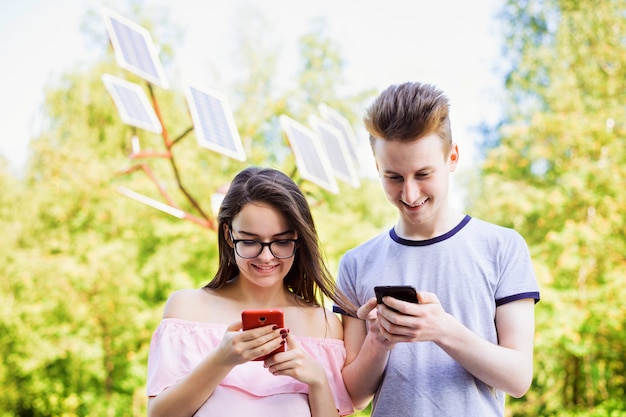 Dwóch wesoło uśmiechniętych studentów korzystających z telefonów komórkowych przed panelami słonecznymi w parku w słoneczny letni dzień