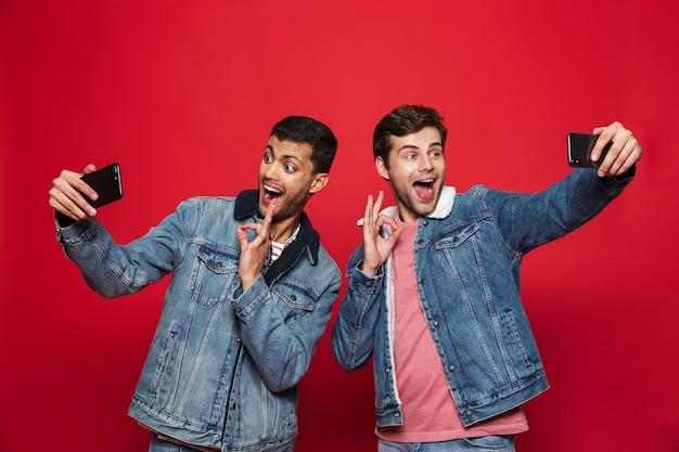 Dwóch wesołych młodych mężczyzn stojących na białym tle nad czerwoną ścianą, biorąc selfie