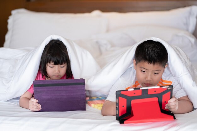 Dwoje chińskich dzieci uzależnionych od tabletu, azjatyckie dziecko oglądające telefon razem na łóżku, dziecko korzystające ze smartfona