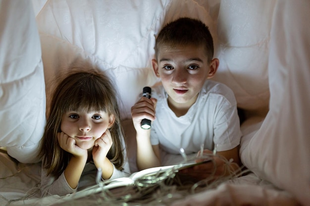 Dwoje dzieci chłopiec i dziewczynka, brat i siostra, przyjaciele leżący pod białym kocem na łóżku, czytający książkę świecącą latarką z lampkami bożonarodzeniowymi patrząc w kamerę uśmiechając się