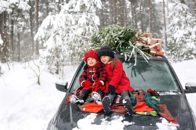 Dwoje dzieci na masce samochodu obok choinki i prezentów