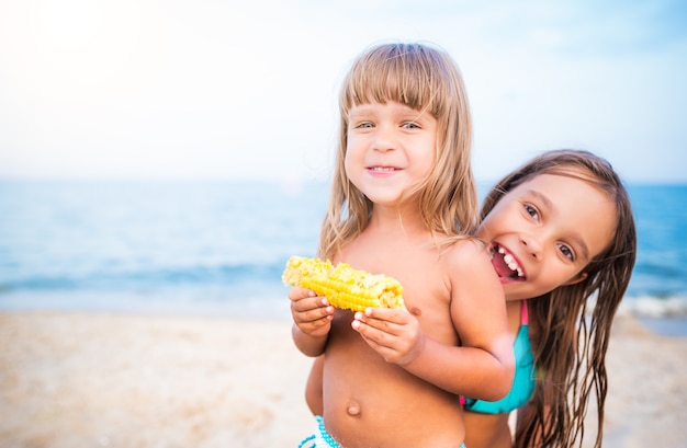 Dwoje Dzieci, Zabawy Na Plaży, Uśmiechnięte I Jedzące Kukurydzę. Dziewczyny Patrząc Na Kamery, Portret Na Rozmytym Tle