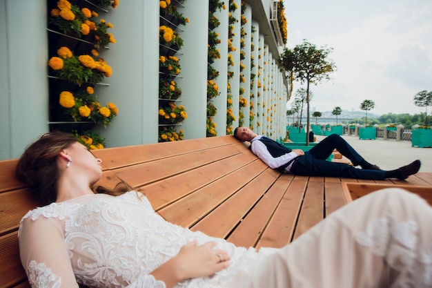 Dwoje kochanków siedzi na ławce, nowożeńcy kucają, by odpocząć w swoich ramionach podczas ślubnej sesji zdjęciowej, panna młoda w białej sukni i pan młody w pięknym garniturze na emeryturze w parku.